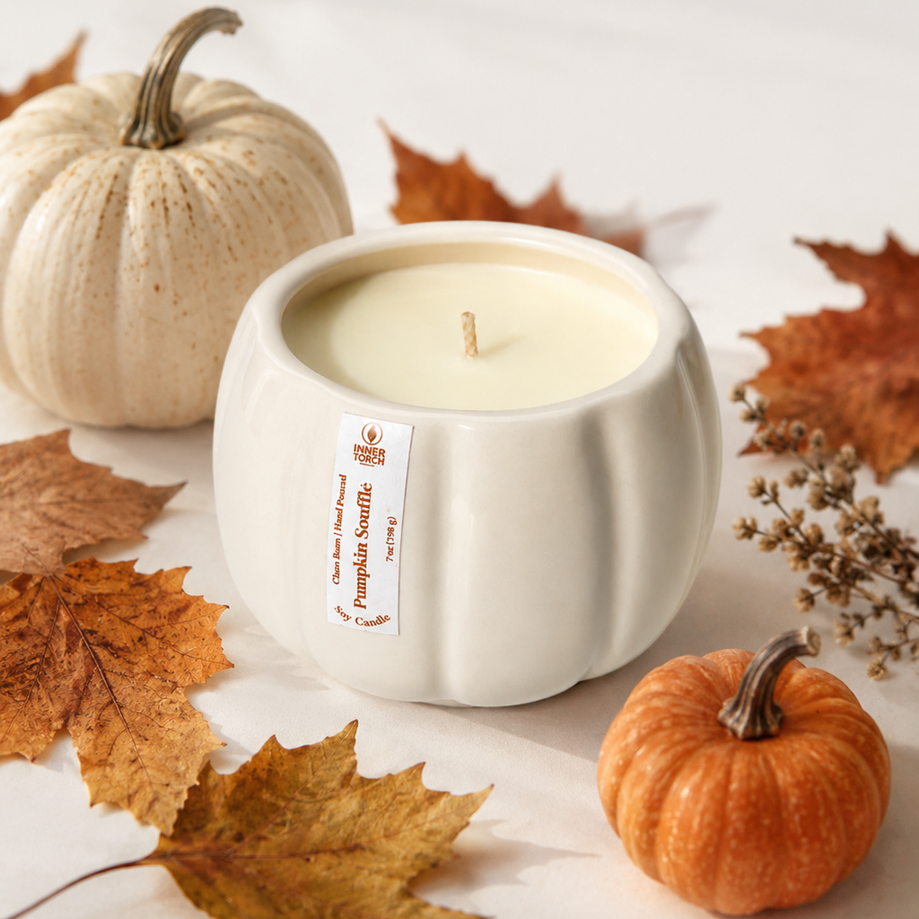 A white ceramic pumpkin-shaped candle with cream wax sits on a light surface surrounded by autumn leaves, a small orange pumpkin, and a larger beige pumpkin. The candle has a visible label reading 'Pumpkin Snuggler' and 'The Art of Scents'.