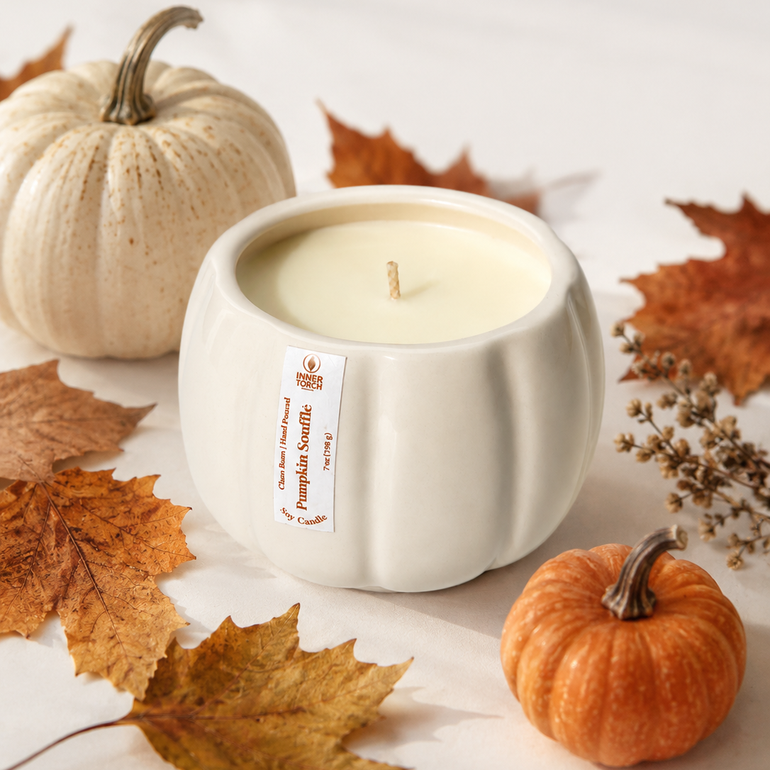 A white ceramic pumpkin-shaped candle with cream wax sits on a light surface surrounded by autumn leaves, a small orange pumpkin, and a larger beige pumpkin. The candle has a visible label reading 'Pumpkin Snuggler' and 'The Art of Scents'.
